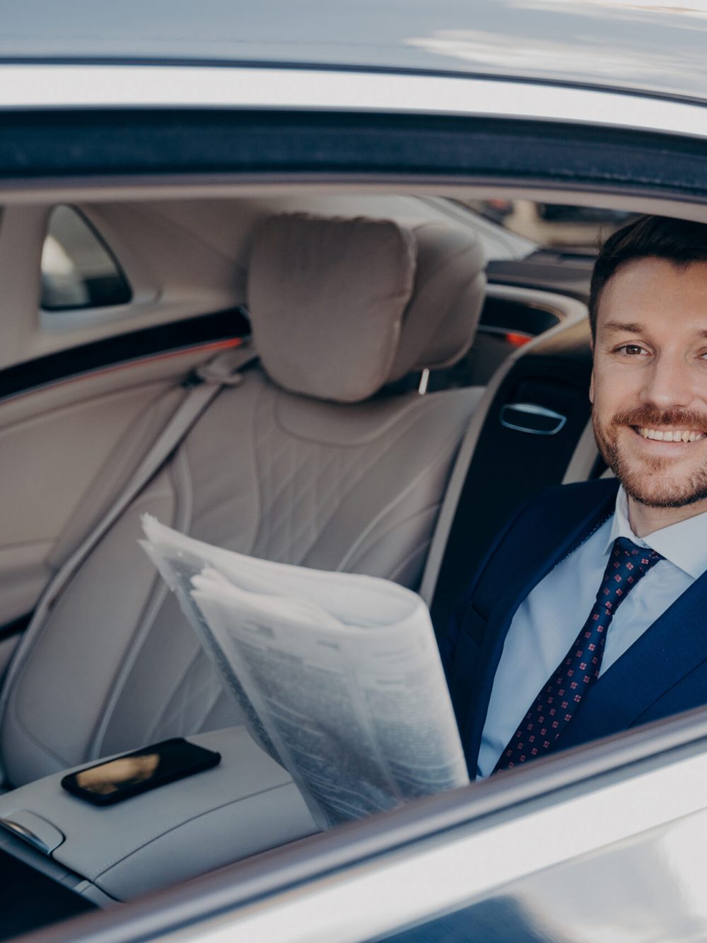 Independent young smiling business owner in blue formal suit, sitting in back of limousine, checking out newspaper, happy to read good news written about himself, taking ride in back of car